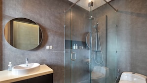 A modern bathroom with a glass-enclosed shower, a round mirror, a white sink, and a toilet. - Aqilah Villa Bali (Legian)