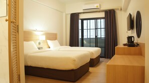 A hotel room with two beds, a wooden headboard, a bedside table with a lamp, a window with curtains, and an air conditioning unit. - Rcn Court (Chiang Mai)