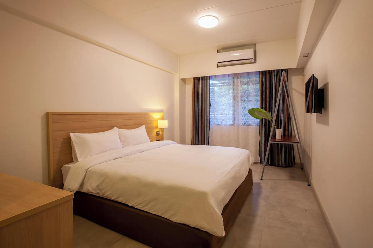 A hotel room with a bed, bedside lamp, air conditioning unit, TV, and a view of the outdoors through a window with curtains.