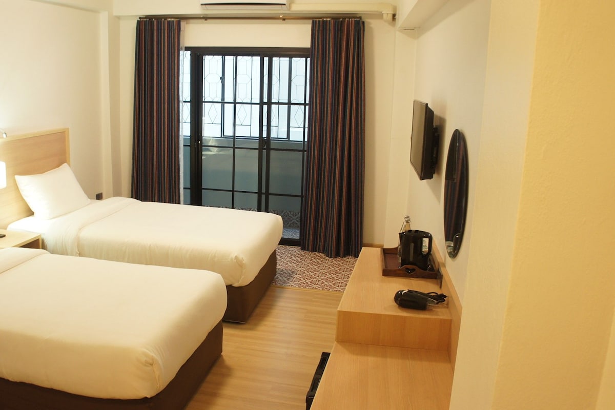 A hotel room with two beds, a small table with a kettle, a TV, and a window with curtains.
