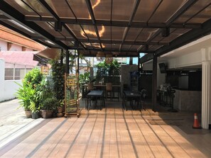 Café al aire libre con mesas y sillas, rodeado de macetas con plantas y un cartel.