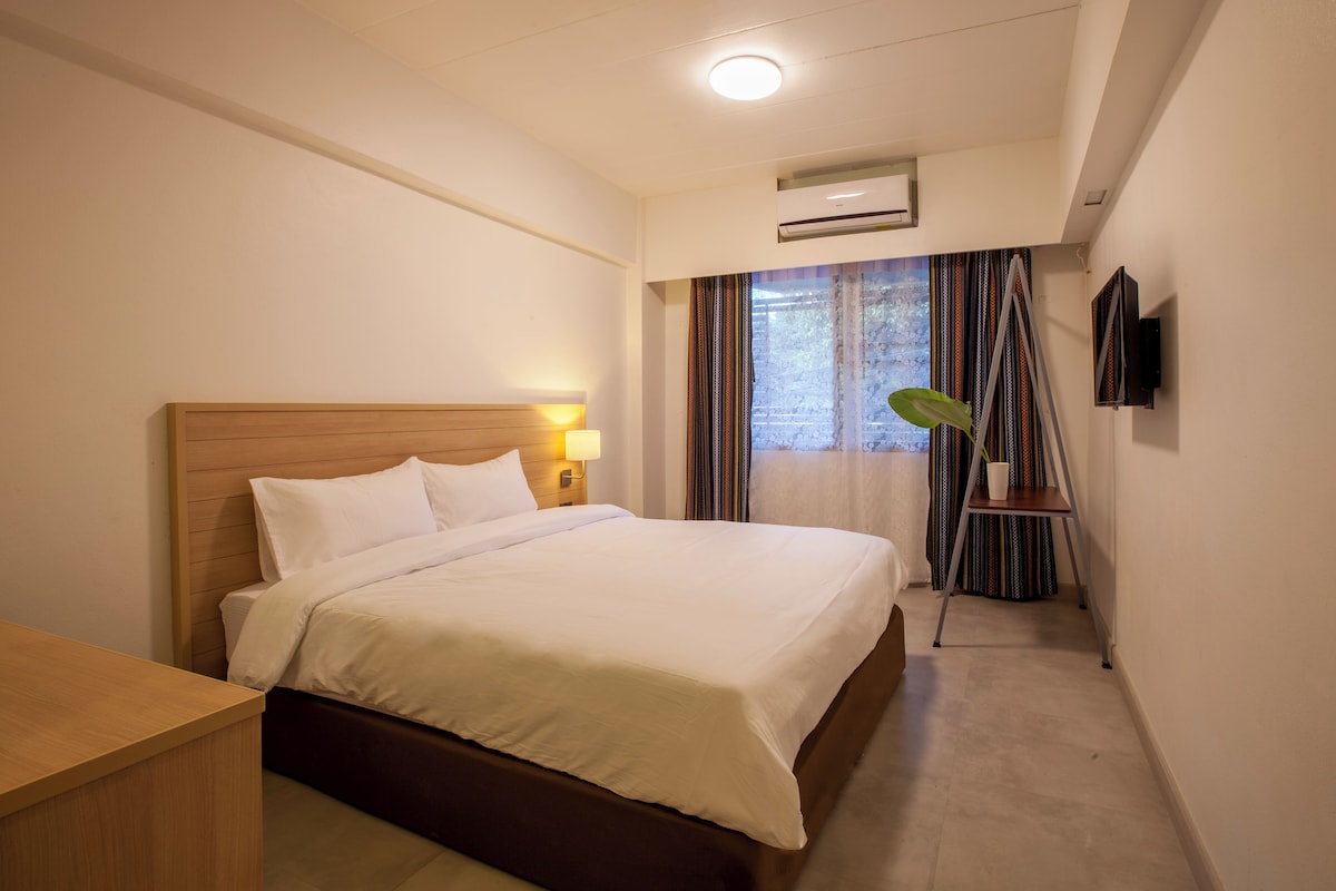 A hotel room with a bed, bedside lamp, air conditioning unit, TV, and a view of the outdoors through a window with curtains.
