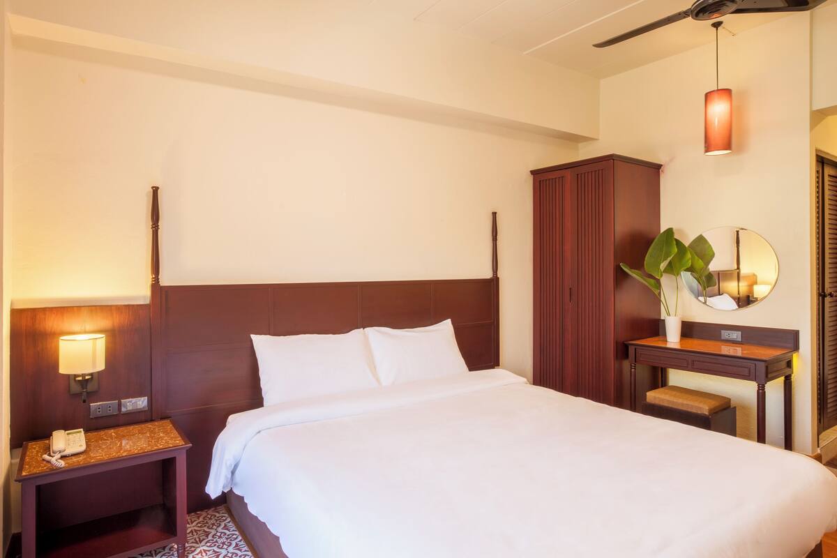 A hotel room with a large bed, a nightstand with a lamp, a wooden wardrobe, a small table with a plant, and a mirror on the wall.