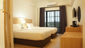 A hotel room with two beds, a wooden headboard, a bedside table with a lamp, a window with curtains, and an air conditioning unit. - Rcn Court (Chiang Mai)