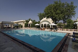 A swimming pool with lounge chairs and umbrellas, surrounded by white buildings with domed structures. - Alsisar Mahal- Heritage Palace Hotel (Malasisar)