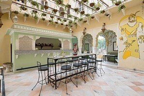 Un comedor con una mesa larga y sillas, una pared decorativa con un mural, plantas colgantes y un estilo arquitectónico tradicional.