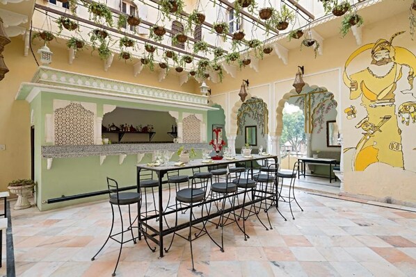A dining area with a long table and chairs, a decorative wall with a mural, hanging plants, and a traditional architectural style. - Alsisar Mahal- Heritage Palace Hotel (Malasisar)