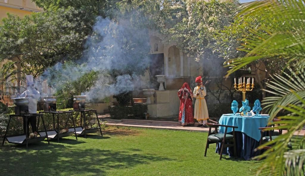 An outdoor dining area with a table set for two, surrounded by greenery and a building in the background.
