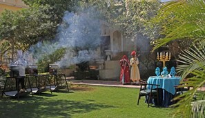 An outdoor dining area with a table set for two, surrounded by greenery and a building in the background. - Alsisar Mahal- Heritage Palace Hotel (Malasisar)