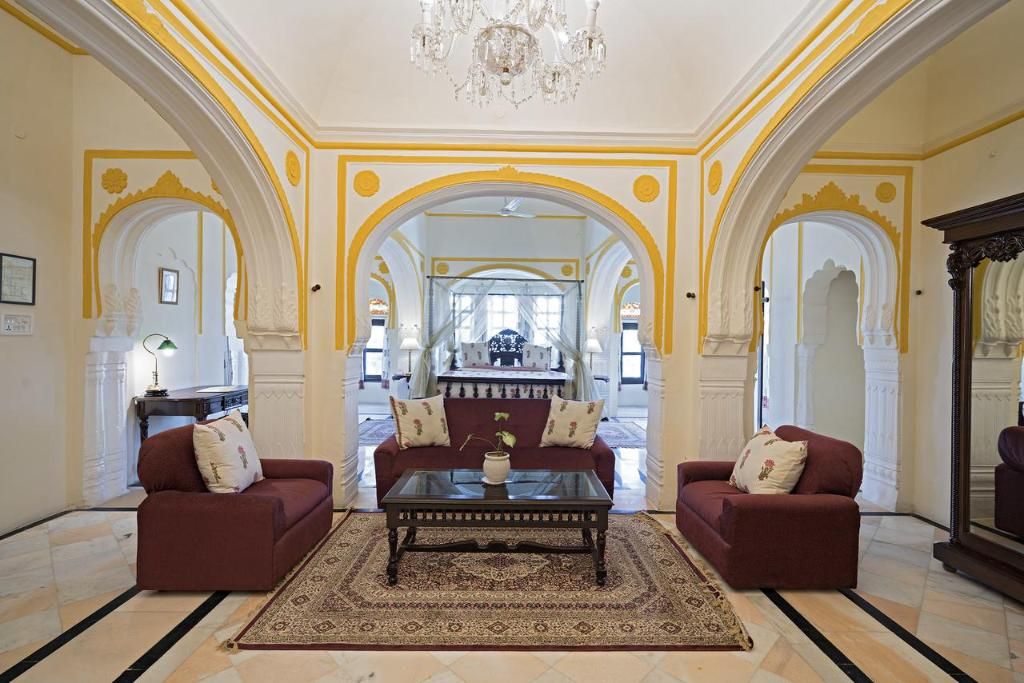 A spacious living room with two red sofas, a central coffee table, and a chandelier. The room features ornate yellow and white arches and a large mirror.