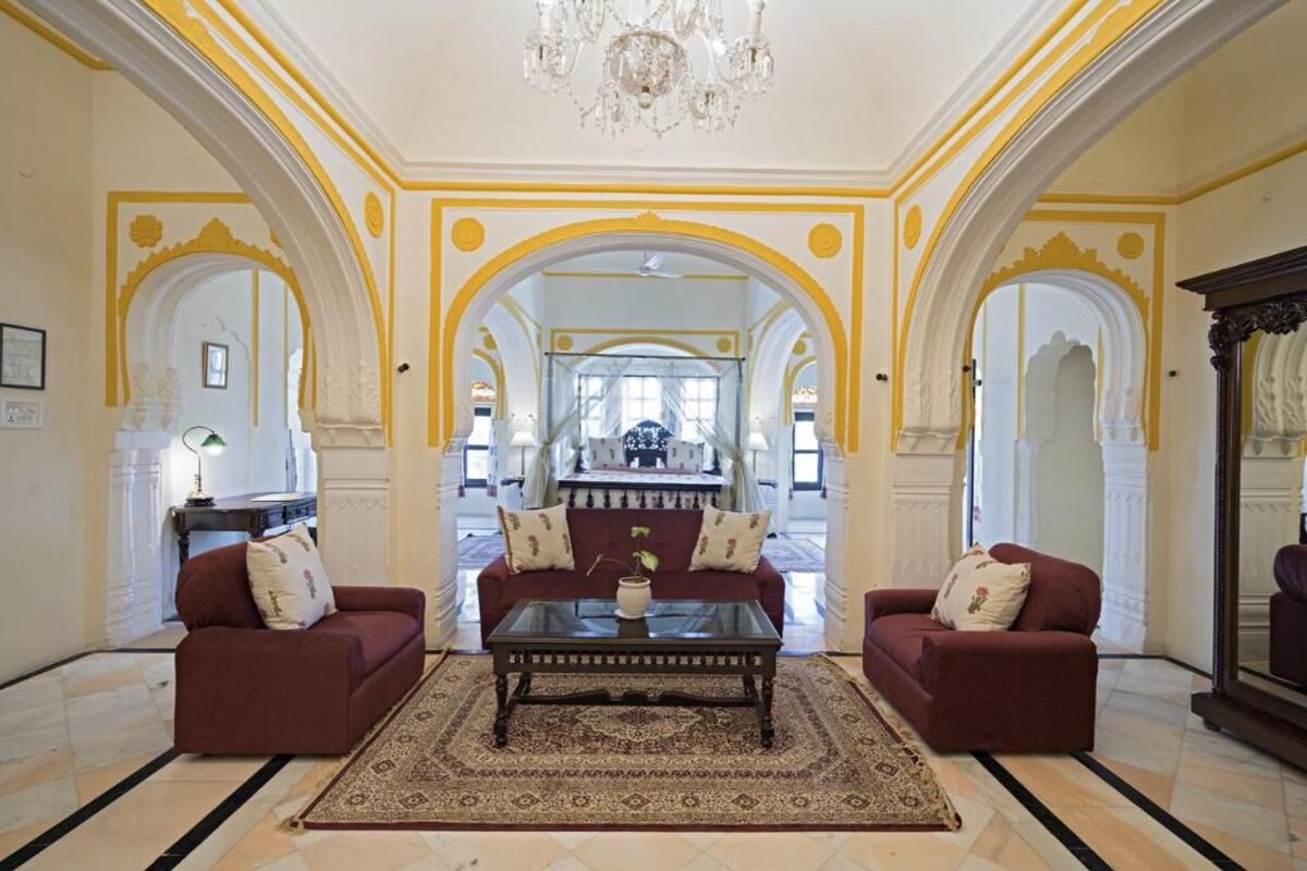 A spacious living room with two red sofas, a central coffee table, and a chandelier. The room features ornate yellow and white arches and a large mirror.