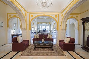 A spacious living room with two red sofas, a central coffee table, and a chandelier. The room features ornate yellow and white arches and a large mirror. - Alsisar Mahal- Heritage Palace Hotel (Malasisar)