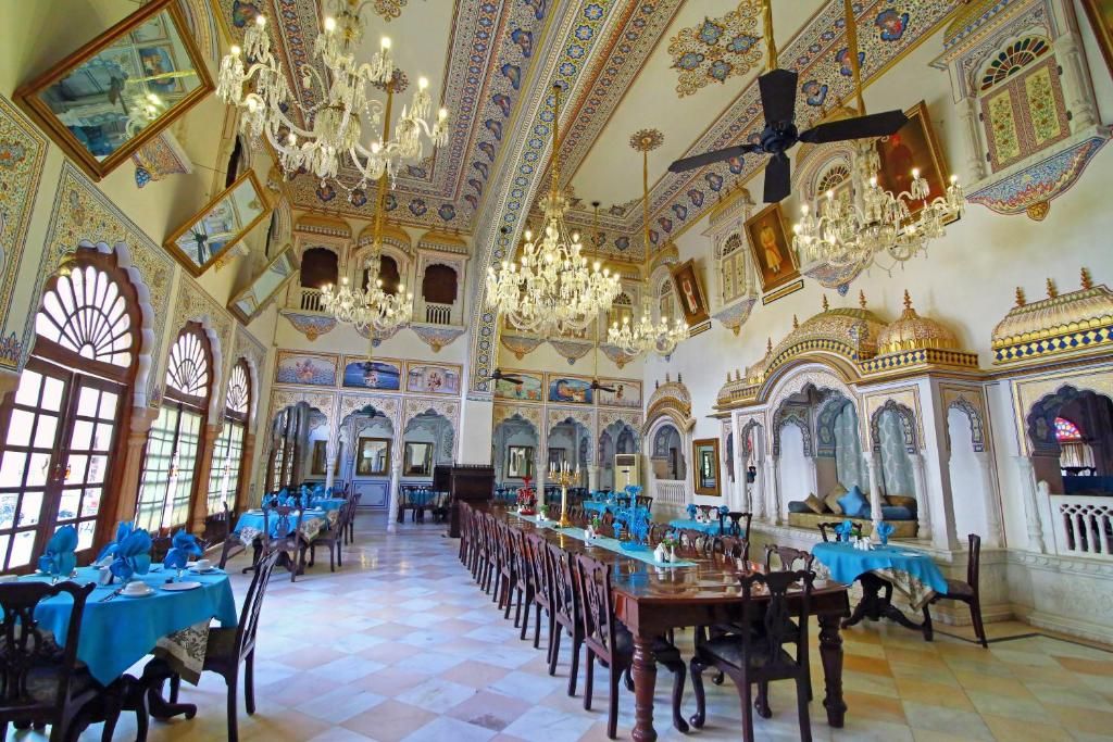 Un salón imponente con candelabros ornamentales, techos altos y diseños intrincados en las paredes. Se han dispuesto mesas para cenar, y la sala cuenta con grandes ventanales.