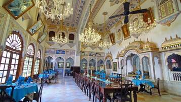 Un salón imponente con candelabros ornamentales, techos altos y diseños intrincados en las paredes. Se han dispuesto mesas para cenar, y la sala cuenta con grandes ventanales.