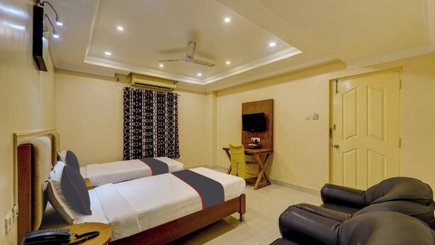 A hotel room with two single beds, a desk with a chair, a sofa, a TV mounted on the wall, and a ceiling fan. - Rg Suites Ashok Nagar (Chennai)