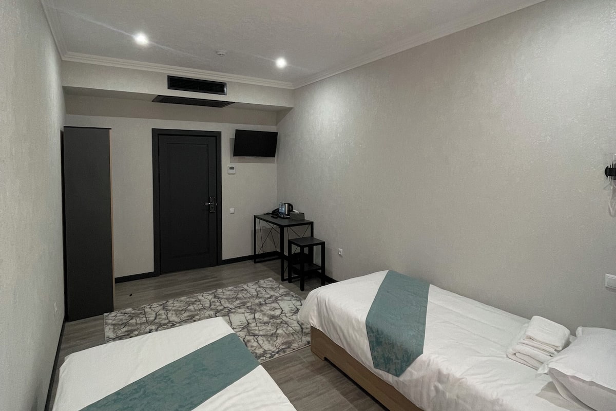 A hotel room with two single beds, a desk with a chair, a small table, and a TV on the wall.