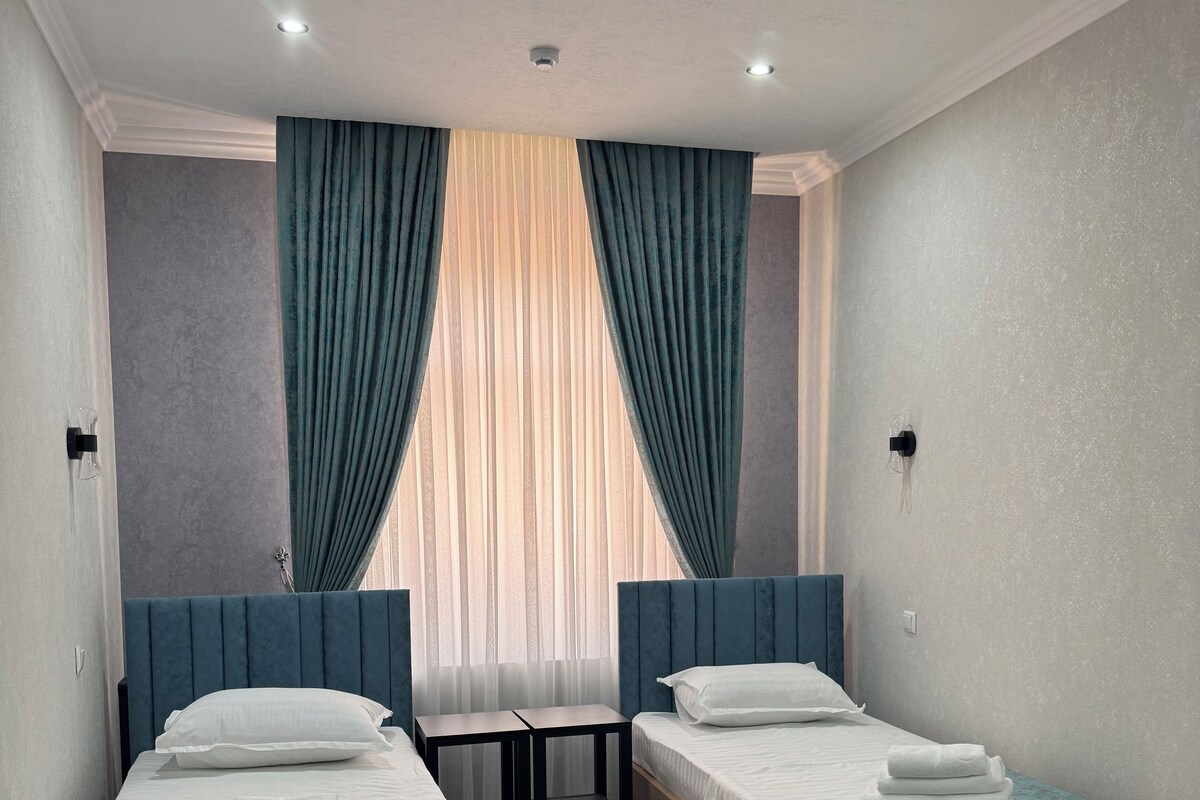 Een hotelkamer met twee enkele bedden, een tafeltje en een raam met gordijnen.