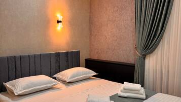 Habitación de hotel con cama doble, dos almohadas, mesita de noche y una lámpara de pared. Hay una ventana con cortinas.