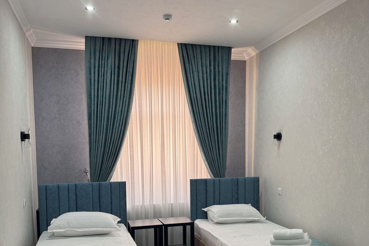 A hotel room with two single beds, a small table, and a window with curtains.