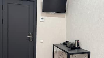 A room with a dark door, a mounted TV, a small table with a phone and a coffee maker, and a trash bin.