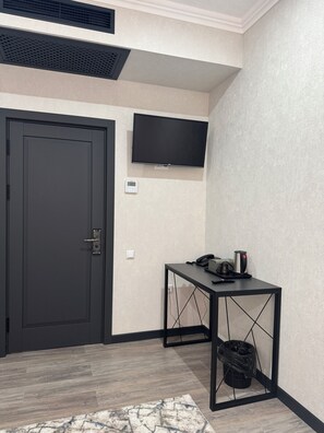 Ein Zimmer mit einer dunklen Tür, einem an der Wand montierten Fernseher, einem kleinen Tisch mit Telefon und Kaffeemaschine sowie einem Mülleimer.