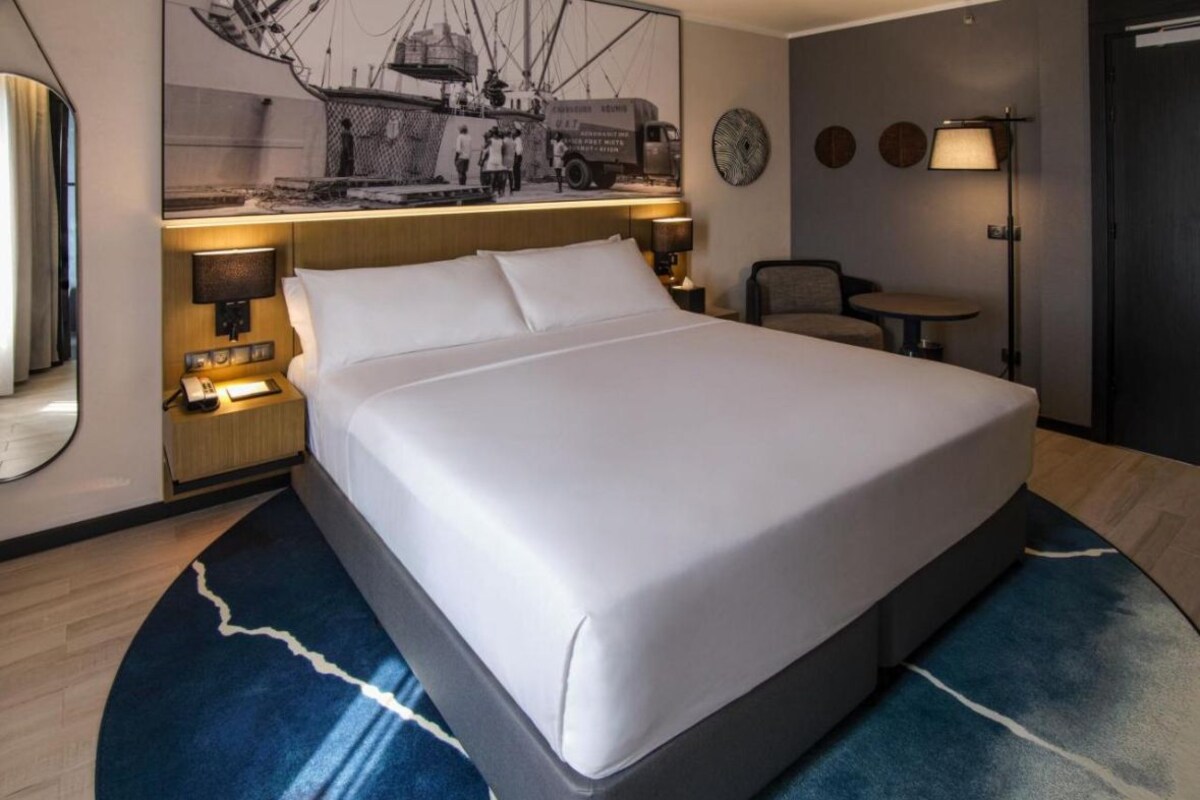 A hotel room with a large bed, bedside lamps, a small table, and a wall-mounted artwork.