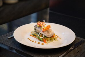 En hvid tallerken med en ret, bestående af kød, grøntsager og pynt, på et mørkt bord.