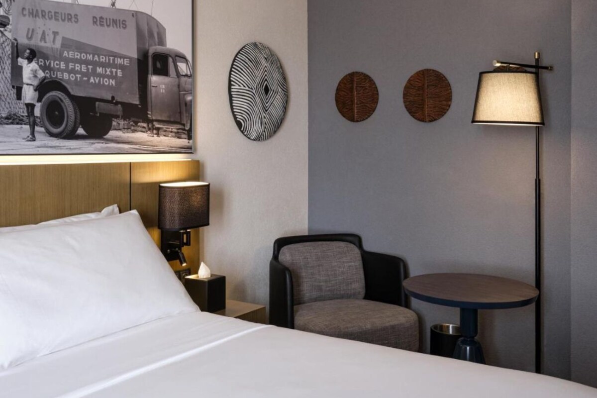 Et hotelværelse med en seng, et natbord med lampe, en stol, et lille bord og vægdekorationer, herunder et plakat og tre runde kunstværker.