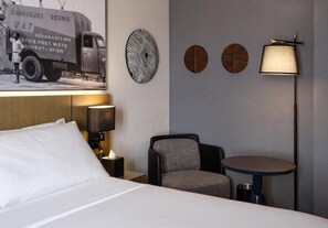 Et hotelværelse med en seng, et natbord med lampe, en stol, et lille bord og vægdekorationer, herunder et plakat og tre runde kunstværker.
