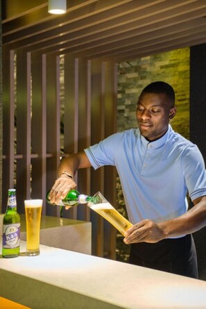 A person pouring beer into a glass at a bar with a bottle and a glass of beer on the counter. - K Hotels (Douala)