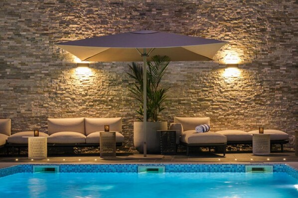 A poolside area with a stone wall, a large umbrella, and lounge chairs. - K Hotels (Douala)