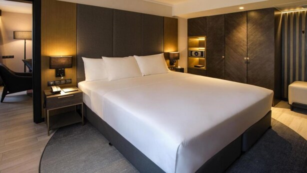 Moderni hotellihuone, jossa on suuri sänky, yöpöytä lampun kanssa, vaatekaappi sekä oleskelualue, jossa on tuoli ja pieni pöytä.