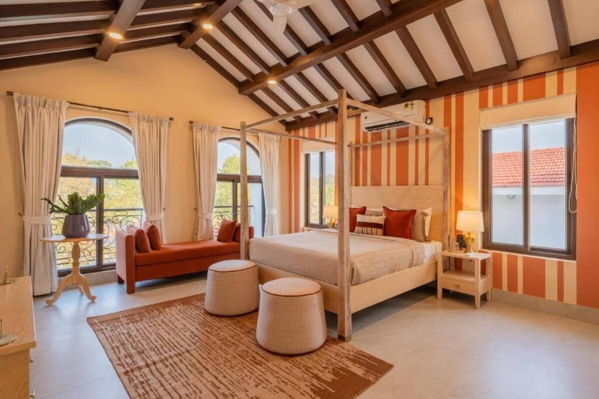 Un dormitorio con una cama con dosel, un sofá, dos pufs y una mesita de noche con lámpara.