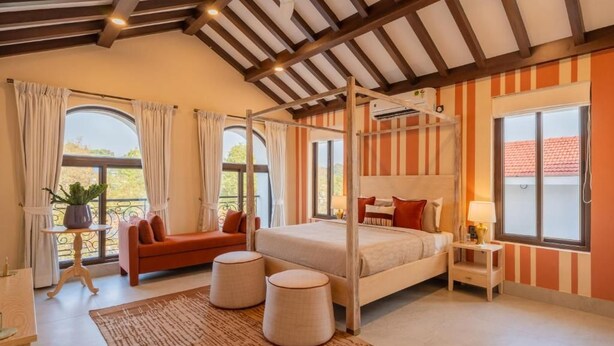 A bedroom with a four-poster bed, a sofa, two ottomans, and a nightstand with a lamp. - Elivaas Paraíso - Villa Goa (Siolim)