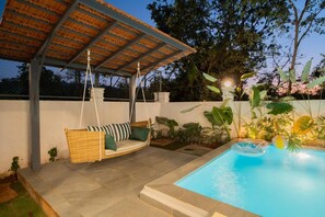 A covered outdoor seating area with a swing chair, a swimming pool, and surrounding greenery. - Elivaas Paraíso - Villa Goa (Siolim)
