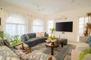 Ein Wohnzimmer mit Ecksofa, Couchtisch, Flachbildfernseher und großem Fenster mit transparenten Vorhängen.