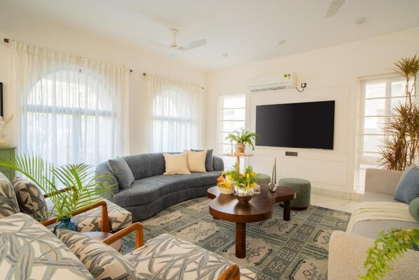 Una sala de estar con un sofá modular, una mesa de centro, un televisor de pantalla plana y un ventanal con cortinas translúcidas.