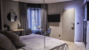 Een moderne hotelkamer met een groot bed, een bureau met stoel, een ronde tafel met stoel, een aan de muur bevestigde televisie en een deur die naar een andere kamer leidt.