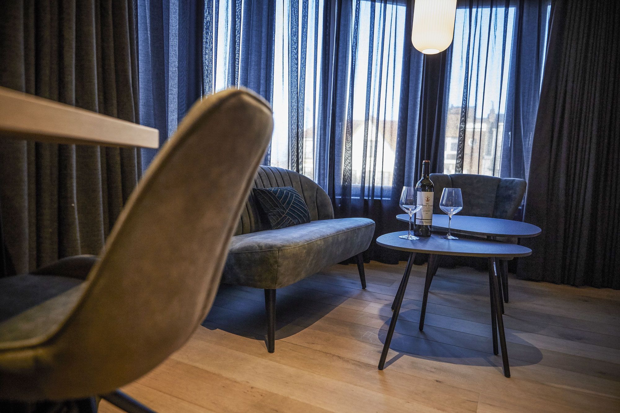 A modern living room with a sofa, a round table with a wine bottle and glass, and a view through large curtains.