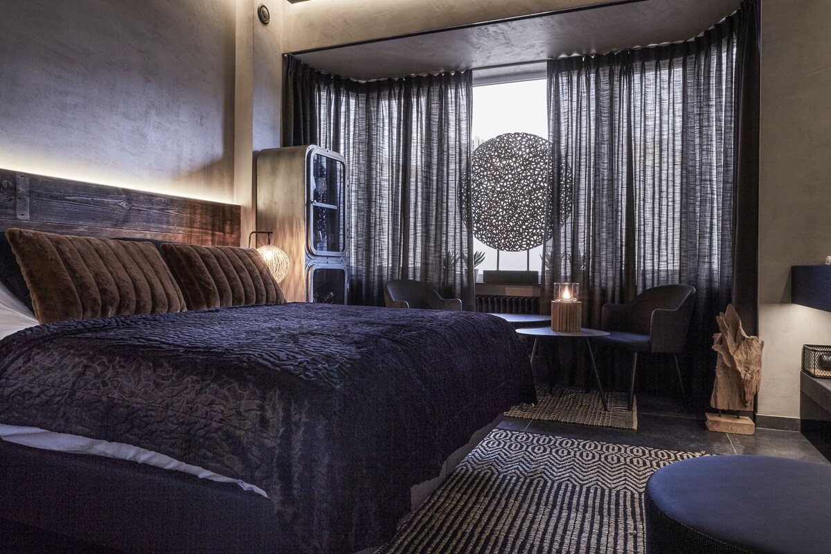 Una camera d'albergo moderna con un letto grande, una scrivania e una sedia. Si vede una finestra con tende e un elemento decorativo a parete.