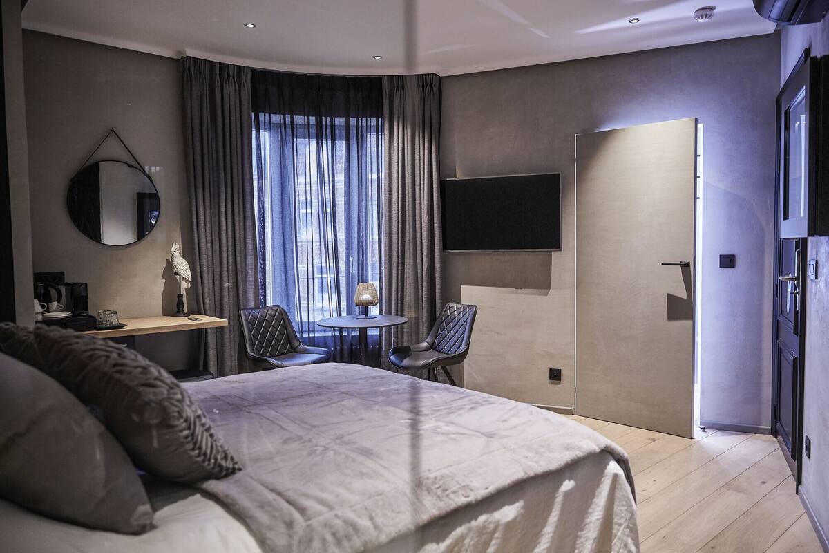 Ein modernes Hotelzimmer mit einem großen Bett, einem Schreibtisch mit Stuhl, einem runden Tisch mit Stuhl, einem an der Wand montierten Fernseher und einer Tür zu einem anderen Raum.