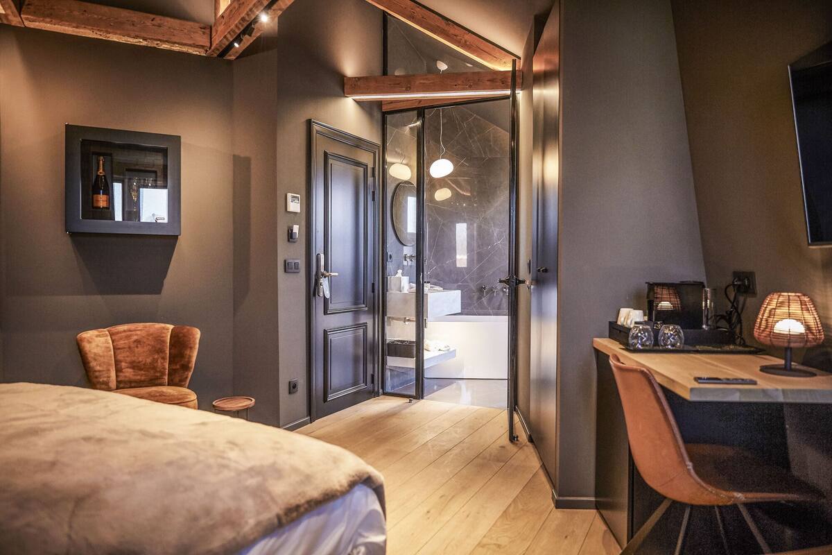 Ein modernes Schlafzimmer mit Holzbalkendecke, einem großen Bett, einem Schreibtisch mit Stuhl und einem durch eine Glastür einsehbaren Badezimmer.
