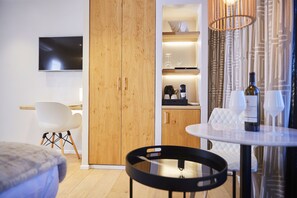 A modern hotel room with a wooden wardrobe, a flat-screen TV, a dining area with a table and chairs, and a kitchen area with a coffee machine and wine bottle. - Hôtel Oniro Tournai - Luxury Rooms & Wellnss Suits (Tournai)