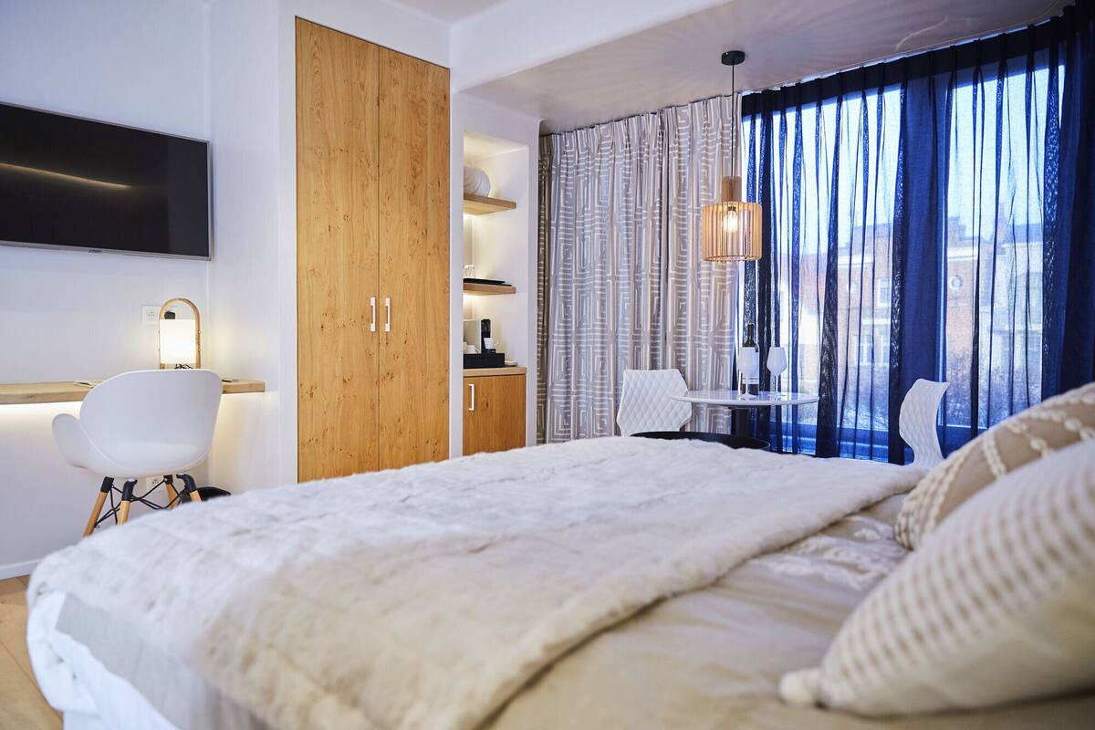 Ein modernes Hotelzimmer mit einem großen Bett, einem Holzschrank, einem Schreibtisch mit Stuhl, einem kleinen Tisch mit Stuhl und einem an der Wand befestigten Fernseher.