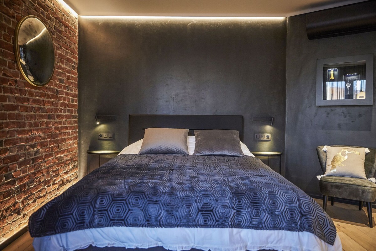 A modern bedroom with a brick wall, a large bed with patterned bedding, two bedside tables with lamps, and a small armchair.