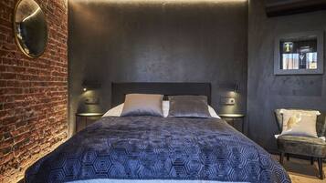 A modern bedroom with a brick wall, a large bed with patterned bedding, two bedside tables with lamps, and a small armchair.