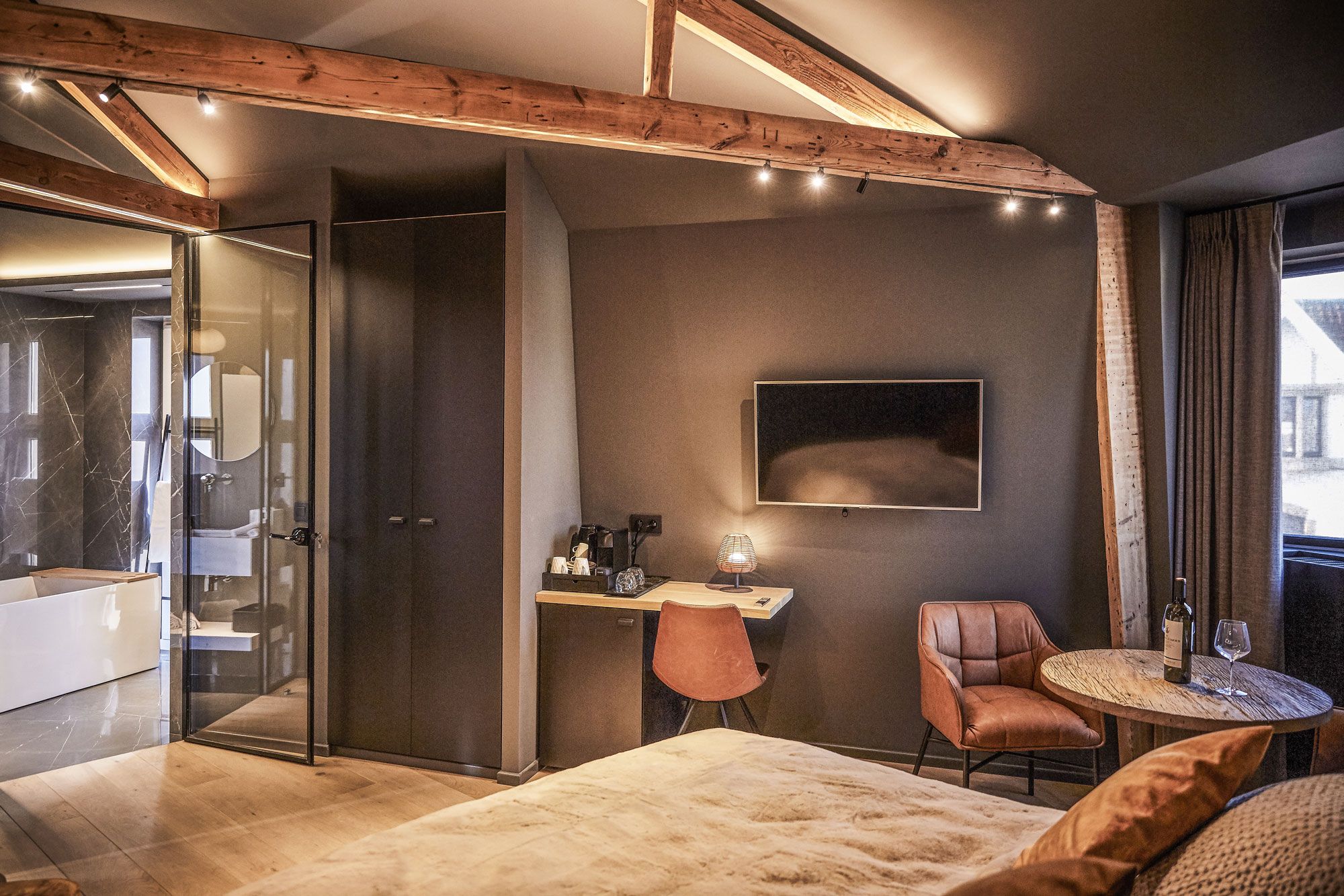 Un dormitorio moderno con ducha de vidrio, techo de madera, una mesa redonda con una copa de vino y una cama grande.
