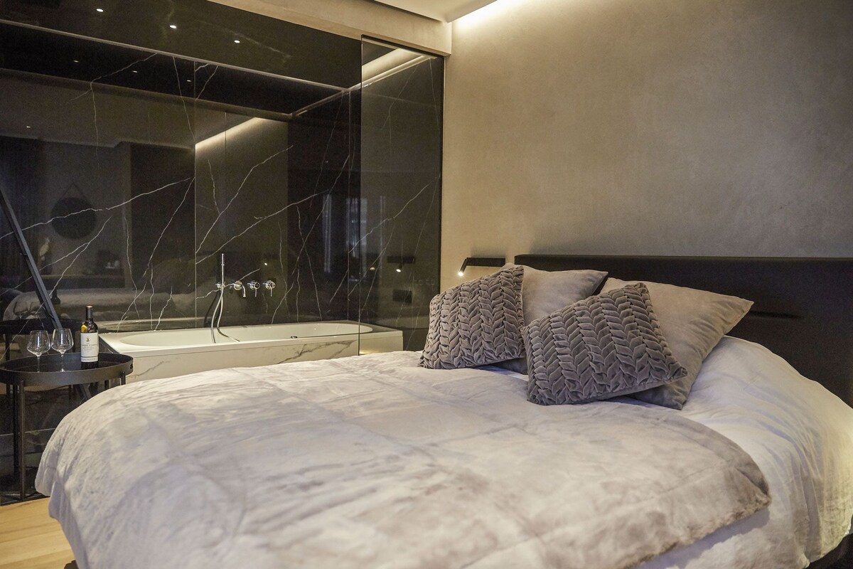 A modern bedroom with a large bed, a glass-enclosed bathtub, and a minimalist design.