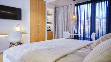 Quarto de hotel moderno com uma cama grande, um armário de madeira, uma escrivaninha com cadeira, uma mesinha com cadeira e uma TV fixada na parede.