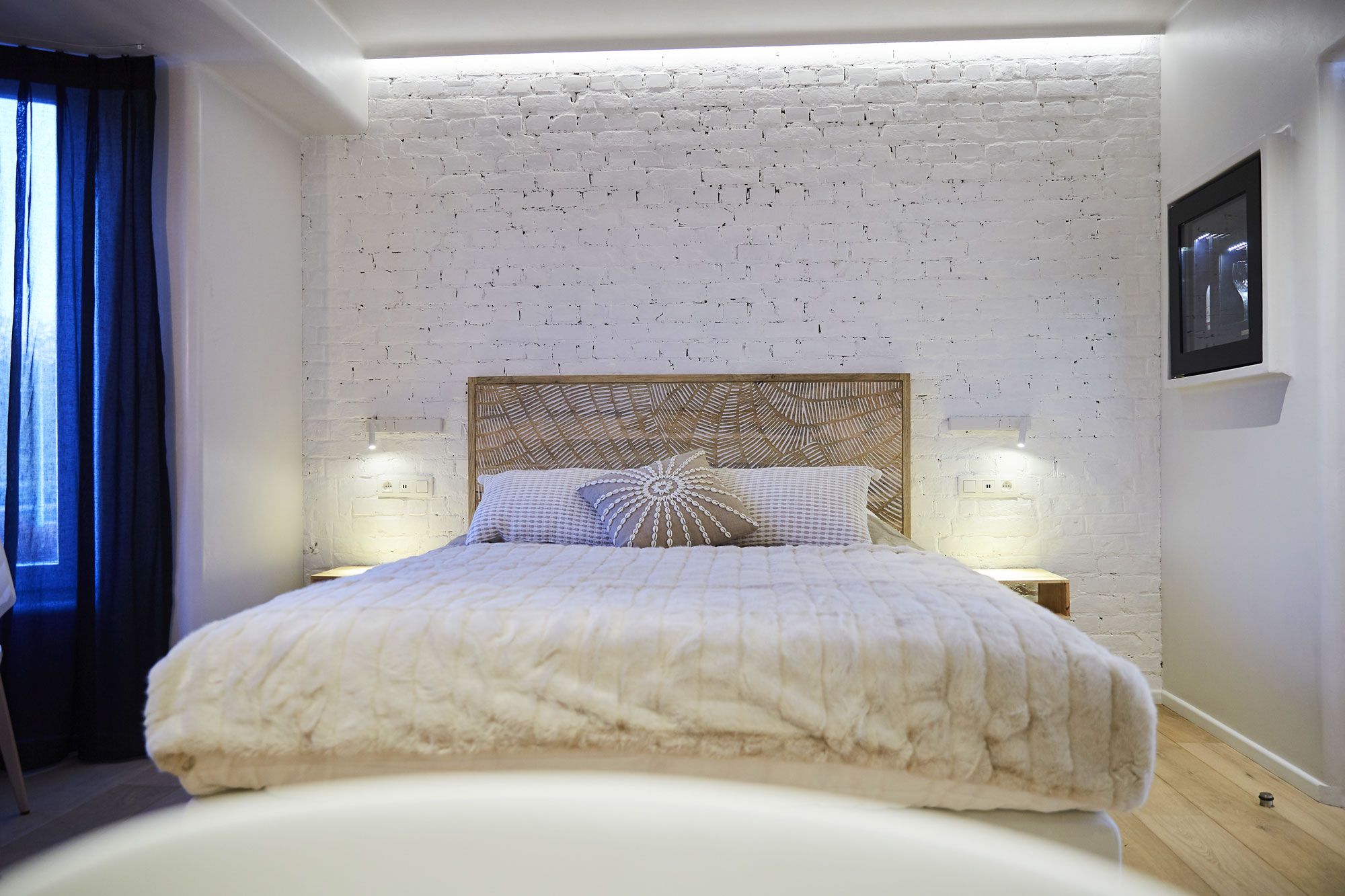 A bedroom with a white brick wall, a wooden headboard, two bedside lamps, and a bed with a white duvet.